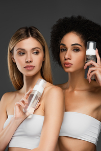 multicultural women in white tops holding bottles with cosmetic foam isolated on grey