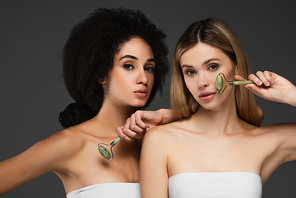 sensual interracial women with perfect skin and natural makeup posing with jade rollers isolated on grey
