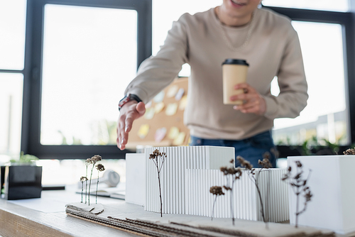 Cropped view of blurred businessman holding paper cup and pointing at model of building in office