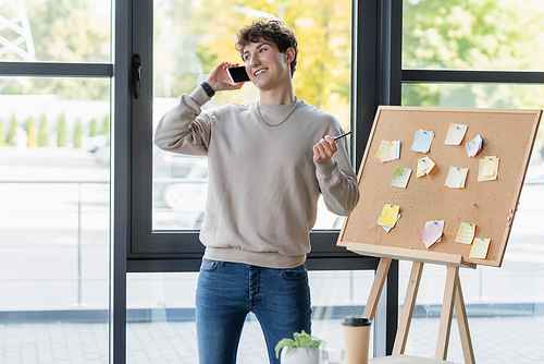 Young businessman talking on smartphone near board with sticky notes in office