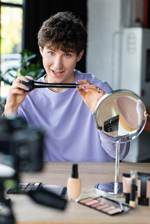 Smiling transgender makeup artist holding cosmetic brush near cosmetics and blurred digital camera