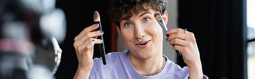 Smiling transgender person holding cosmetic brushes near digital camera, banner