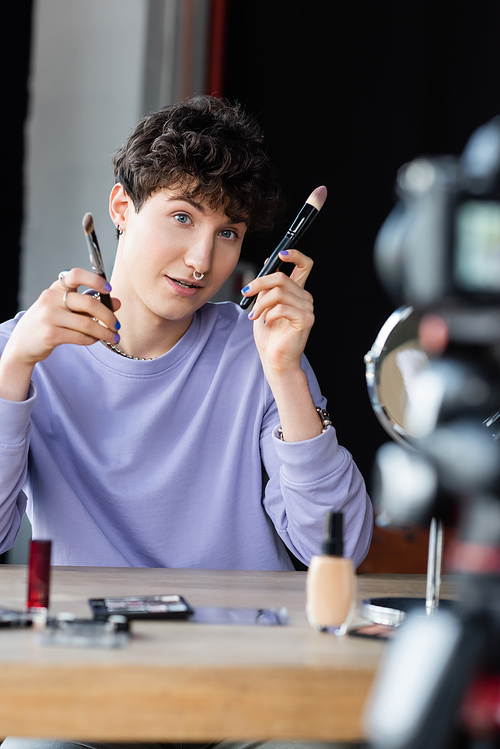 Transgender makeup artist holding cosmetic brushes near blurred digital camera