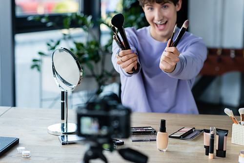 Blurred transgender makeup artist holding cosmetic brushes near digital camera in studio