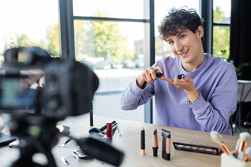 Smiling transgender person holding cosmetics near digital camera
