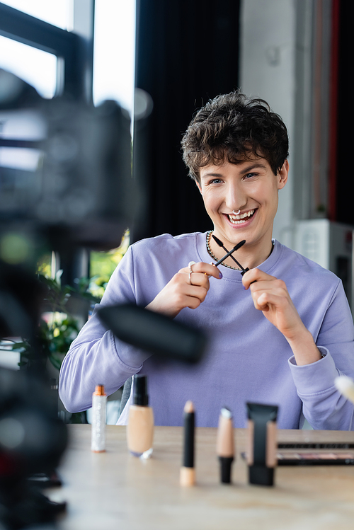 Smiling transgender person holding mascara brushes near decorative cosmetics and digital camera