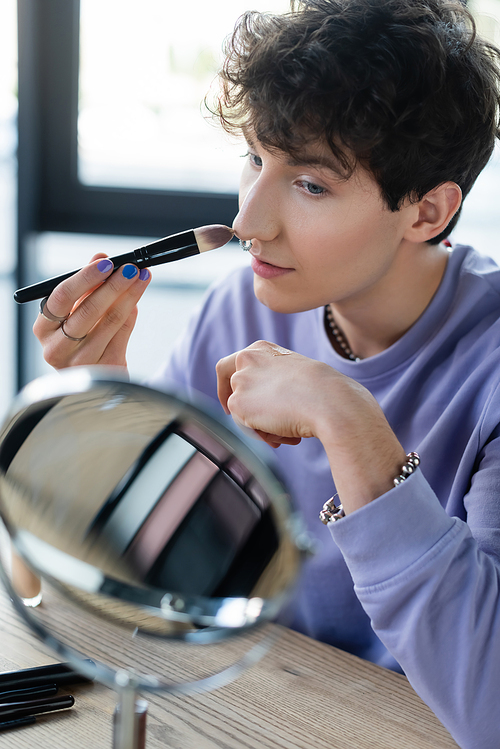 Young transgender person applying makeup foundation near mirror in studio