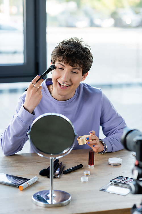 Smiling transgender person applying face powder near mirror, decorative cosmetics and digital camera