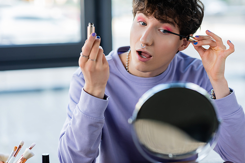 Transgender person applying mascara near cosmetic brushes and mirror