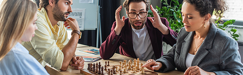 Astonished businessman sitting near interracial colleagues playing chess in office, banner