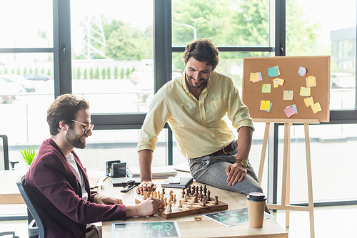 Smiling businessmen playing chess near coffee to go in office
