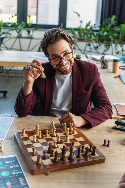 Smiling businessman in eyeglasses holding chess figure near board in office