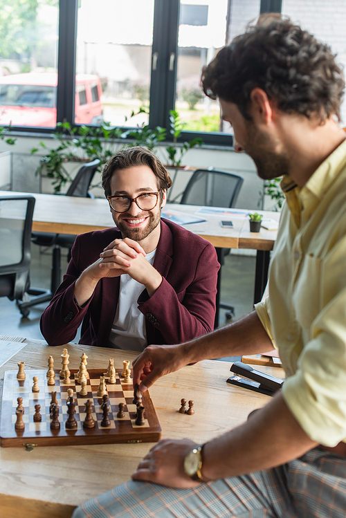 Smiling businessman in eyeglasses looking at blurred colleague playing chess in office