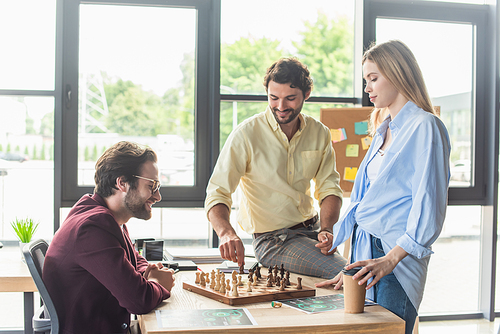 Smiling business people playing chess near colleague with coffee to go in office