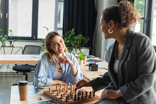 Smiling businesswoman looking at african american colleague playing chess near papers and coffee in office