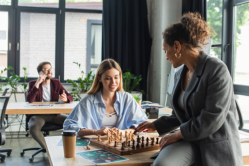 Interracial businesswomen playing chess near blurred colleague in office