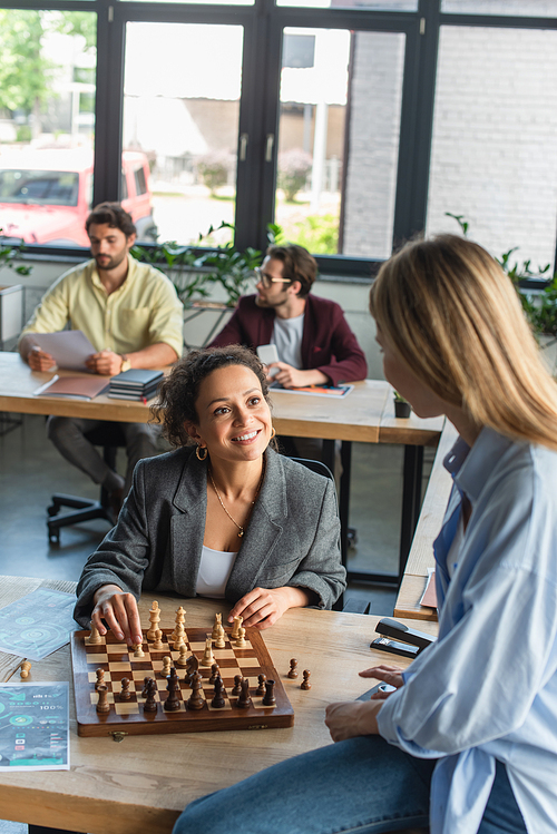 Interracial businesswomen playing chess near blurred businessmen in office