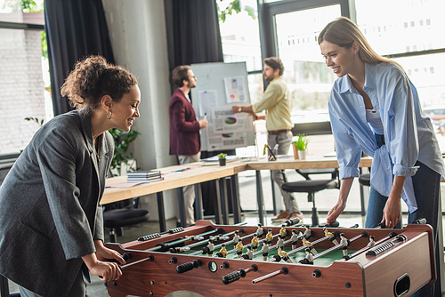 Cheerful interracial businesswomen playing table soccer while blurred colleagues working in office