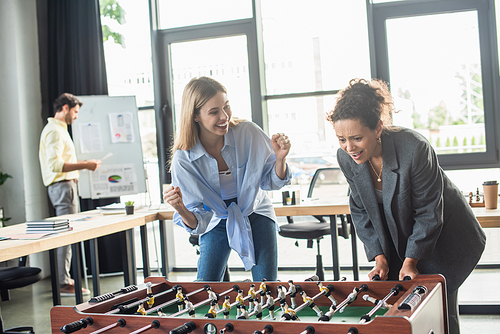 Excited businesswoman showing yes gesture near african american colleague playing table soccer in office