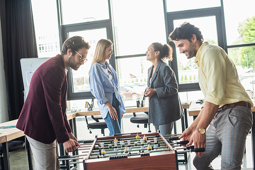 Businessmen playing table soccer near interracial colleagues talking in office