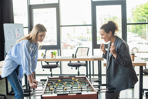 Excited african american businesswoman showing yes gesture while playing table soccer with colleague in office