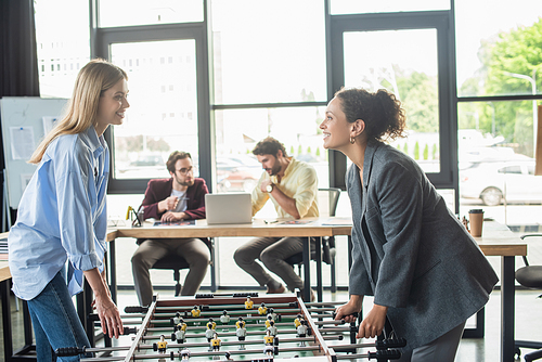 Cheerful businesswomen playing table soccer near blurred businessmen in office