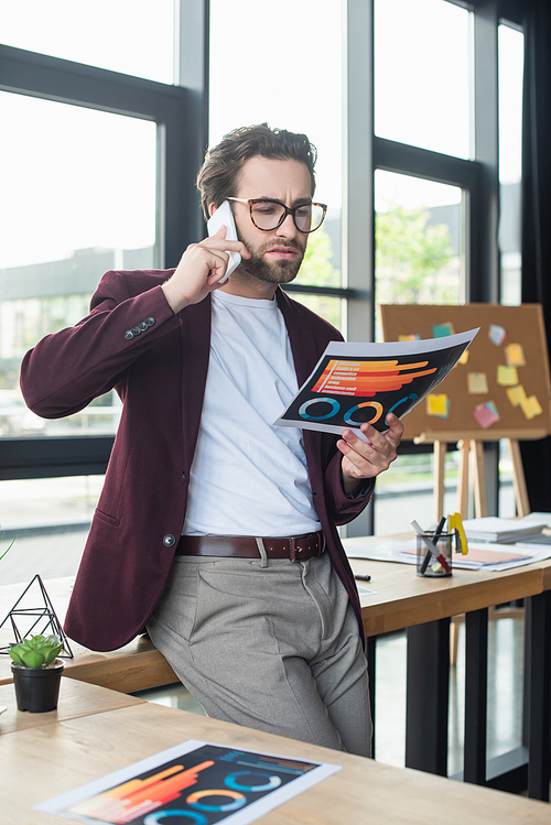 Businessman in eyeglasses talking on smartphone and holding document in office