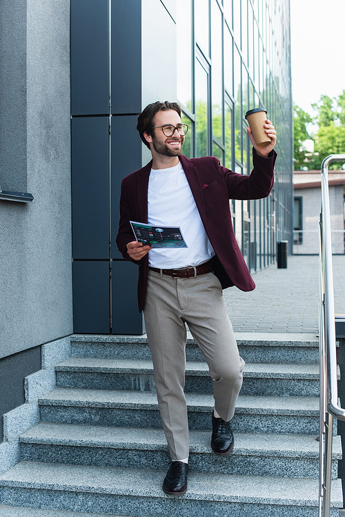 Cheerful businessman with paper cup and document walking on stairs near building