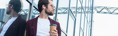 Side view of businessman in eyeglasses holding coffee to go near building on urban street, banner
