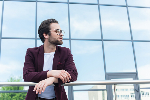 Young businessman in eyeglasses standing near railing and building on urban street