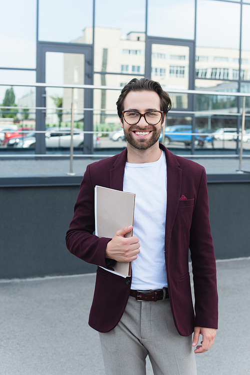 Businessman in formal wear holding paper folders and smiling at camera outdoors