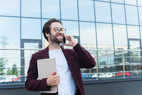Positive businessman with paper folders talking on smartphone on urban street