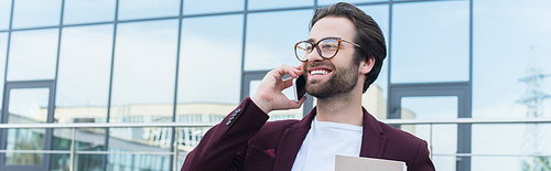 Young businessman with paper folder smiling while talking on smartphone outdoors, banner
