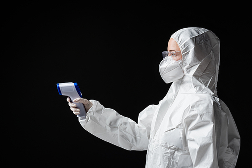 side view of doctor in hazmat suit, goggles and medical mask holding pyrometer isolated on black