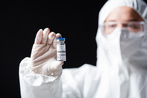 blurred immunologist in white hazmat suit holding covid-19 omicron variant vaccine isolated on black