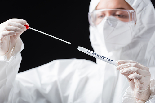 blurred scientist in hazmat suit holding covid-19 omicron variant swab test isolated on black