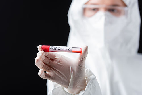 selective focus of positive covid-19 omicron variant test in hand of blurred microbiologist in hazmat suit isolated on black