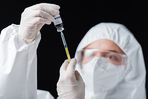 blurred immunologist in hazmat suit holding syringe and covid-19 omicron variant vaccine isolated on black