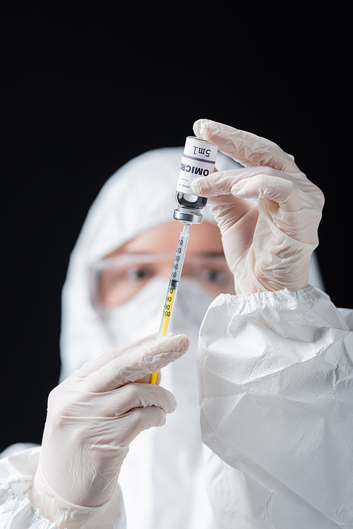 blurred immunologist in hazmat suit holding syringe and omicron variant vaccine isolated on black