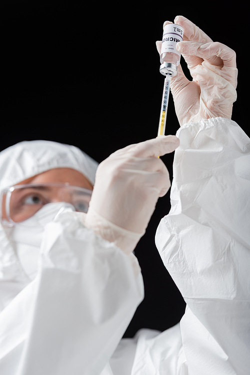 blurred doctor in personal protective equipment holding syringe and omicron variant vaccine isolated on black