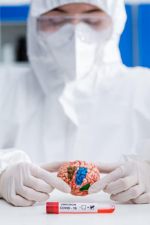 blurred doctor in hazmat suit holding brain model near positive covid-19 omicron variant test