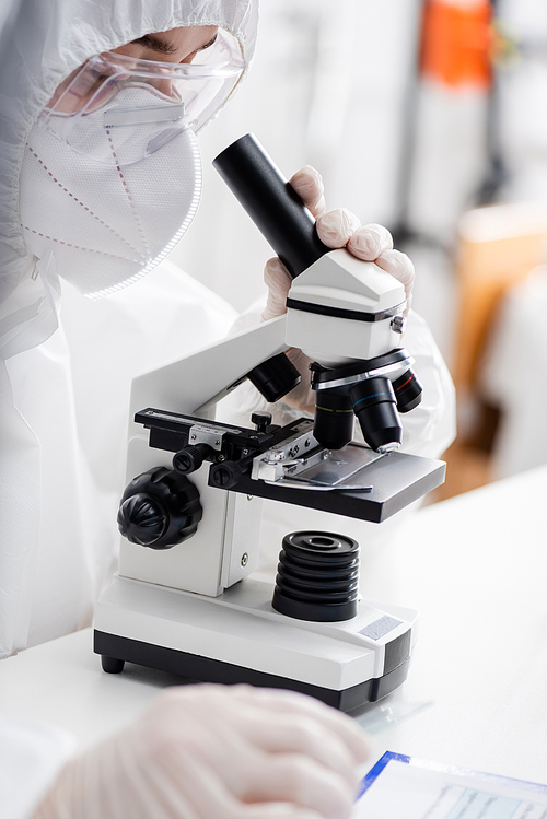 biologist in hazmat suit, goggles and medical mask working with microscope in lab, omicron variant concept