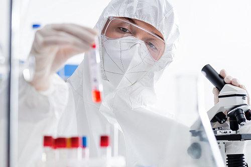 scientist in goggles, medical mask and hazmat suit holding blurred test tube near microscope in lab