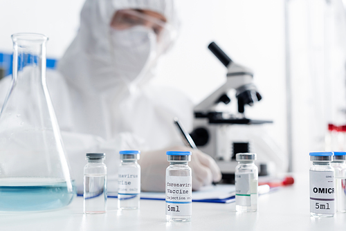 selective focus of vials with coronavirus vaccine near scientist writing near microscope on blurred background