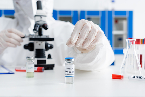 cropped view of blurred scientist working with microscope near coronavirus vaccine and test tubes