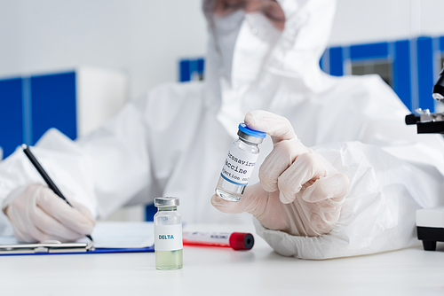 blurred doctor in hazmat suit holding coronavirus vaccine while writing on clipboard in laboratory