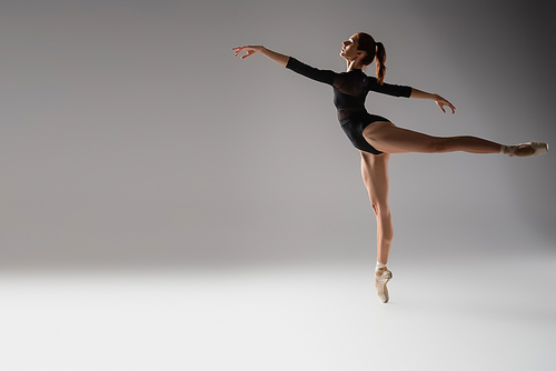 full length of woman in ballet shoes dancing with outstretched hands on dark grey