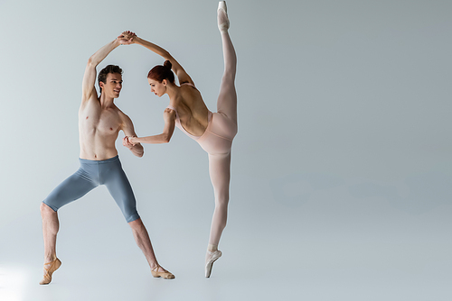 full length of young cheerful ballet dancer performing ballet dance with ballerina on grey