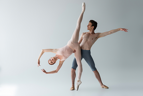 shirtless ballet dancer supporting flexible ballerina with outstretched hands isolated on grey
