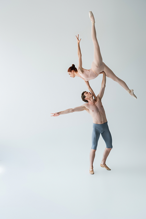 full length of shirtless ballet dancer lifting graceful ballerina in bodysuit on gray